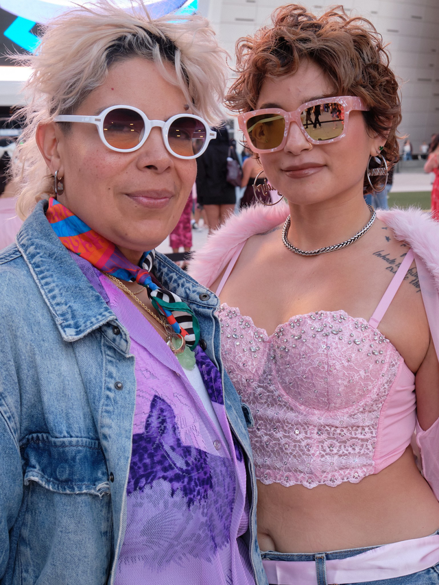 Two people wearing sunglasses pose closely; one wears a denim jacket and colorful scarf, the other a pink beaded crop top and chain necklace.